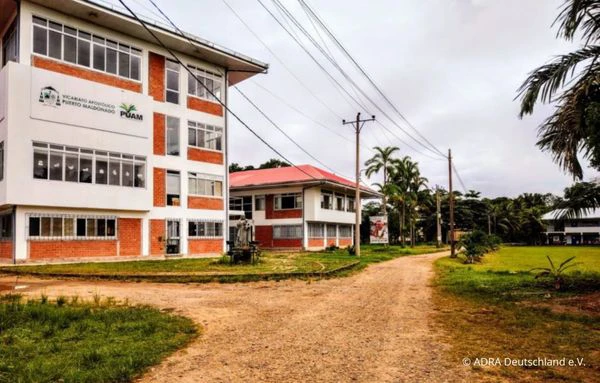 Einfach eingerichtetes Einzelzimmer auf dem Universitätsgelände mit Bett, Schreibtisch, Stuhl, Regal und Ventilator. Durch ein Fenster mit Vorhängen fällt Tageslicht in den Raum.