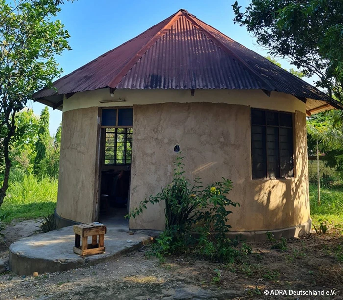 Rundes, einfaches Haus mit Blechdach auf dem Gelände von „The Baobab Home“ in Bagamoyo, umgeben von Bäumen und Grünflächen.