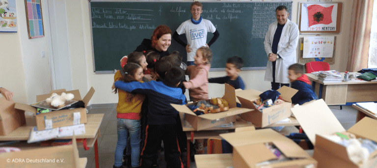 Ein ADRAlive!-Freiwilliger hilft in einer albanischen Schulklasse bei der Verteilung von Paketen im Rahmen der Aktion 'Kinder helfen Kindern!'. Die Kinder sind aufgeregt und umarmen sich gegenseitig. Im Hintergrund sind die Pakete auf den Schultischen zu sehen.