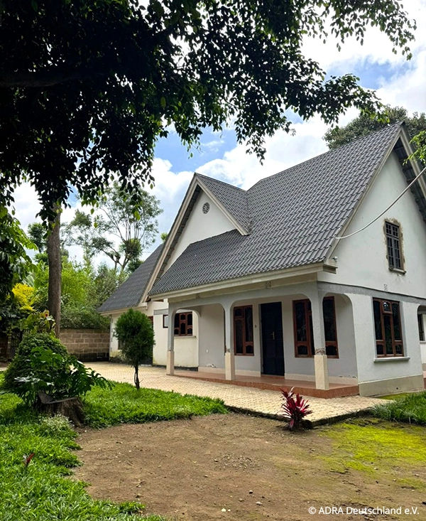 Helle Freiwilligenunterkunft in Arusha, Tansania mit steilem grauen Dach, überdachter Veranda und kleinem Garten, umgeben von Bäumen und Grünflächen