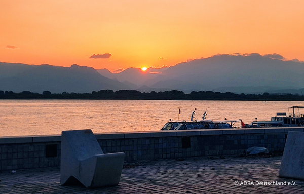 Sonnenuntergang über einem Fluss mit Bergsilhouette in Albanien. Boote am Ufer und orangefarbener Himmel.