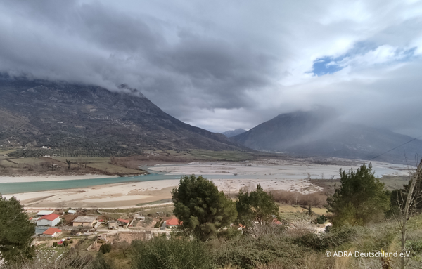 Albanies Flusslandschaft in einem Tal, umgeben von hohen Bergen und kleinen Häusern im Vordergrund, Wolken hängen tief über den Gipfeln.