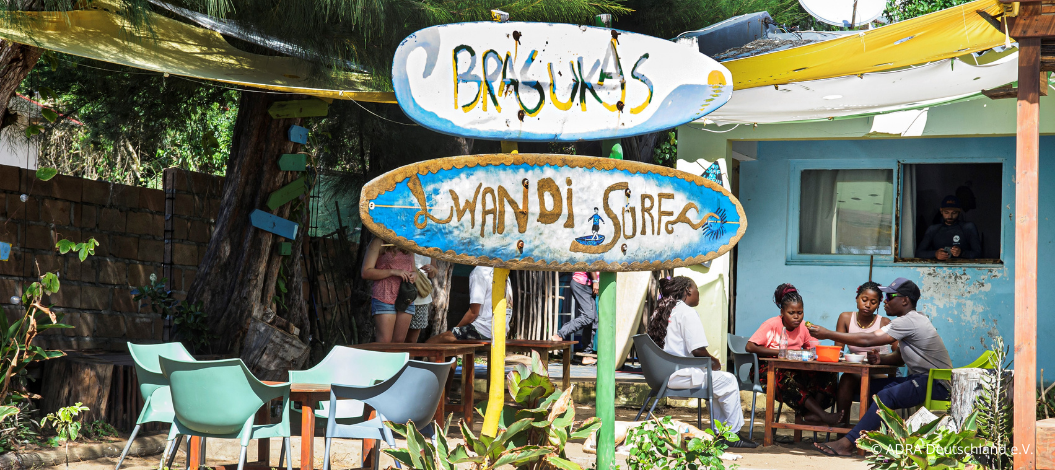 Handgemaltes Surfschild „Lwandi Surf“ vor einem kleinen Café mit Menschen, die draußen an einem Tisch sitzen.
