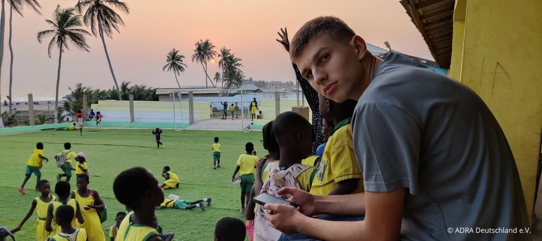 Samuel sitzt am Spielfeldrand und beobachtet Kinder in gelben und grünen Trikots, die auf einem Fußballplatz spielen. Im Hintergrund geht die Sonne über dem Meer unter.