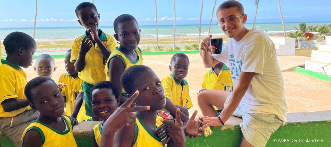 Simon sitzt mit mehreren lachenden Schulkindern in gelben Uniformen auf einer Mauer. Einige Kinder posieren fröhlich für die Kamera, im Hintergrund sind Palmen und das Meer sichtbar.