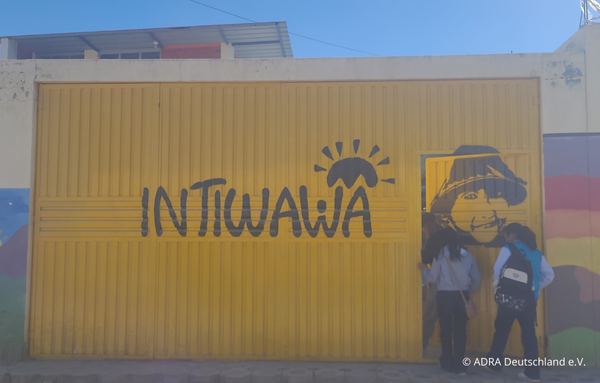Gelbes Metalltor mit großem schwarzen ‚INTIWAWA‘-Schriftzug und Sonnenlogo; seitliche Tür offen, zwei Jugendliche treten ein.