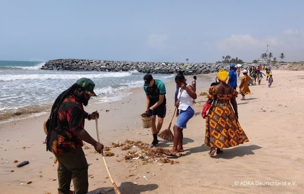 Mehrere Erwachsene und Jugendliche kehren und sammeln Müll am Strand, während Wellen im Hintergrund an die Küste schlagen.