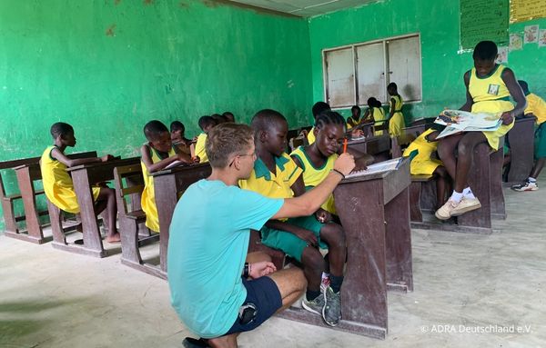 Freiwilliger kniet neben einem Schüler in einem Klassenzimmer mit grüner Wand und hilft ihm beim Schreiben. Mehrere Kinder in gelben Uniformen sitzen an Holzbänken und arbeiten an ihren Aufgaben.