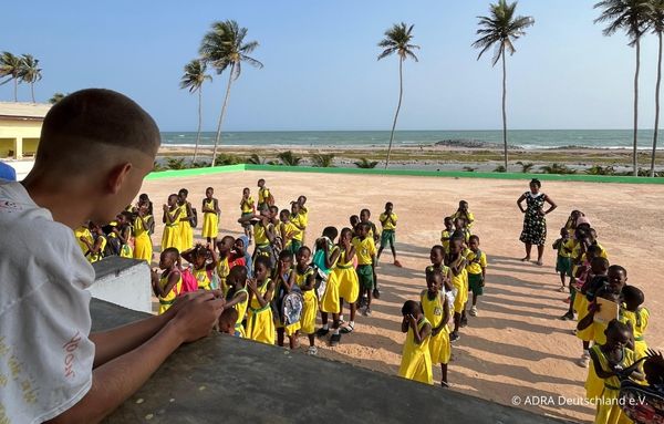 Simon blickt von einem Balkon auf eine große Gruppe Schulkinder in gelben Uniformen, die sich im Schulhof versammeln. Im Hintergrund sind Palmen und das Meer zu sehen.