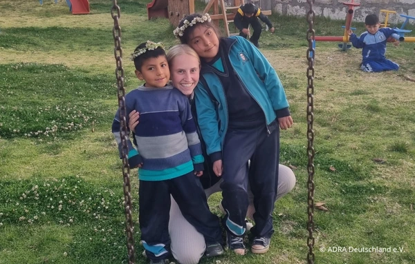 Eine ADRAlive!-Freiwillige kniet zwischen zwei Kindern auf einem Spielplatz und umarmt sie lächelnd.
