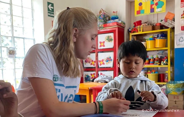 Eine ADRAlive!-Freiwillige sitzt mit einem kleinen Jungen an einem Tisch und bastelt gemeinsam in einem bunten Klassenraum.