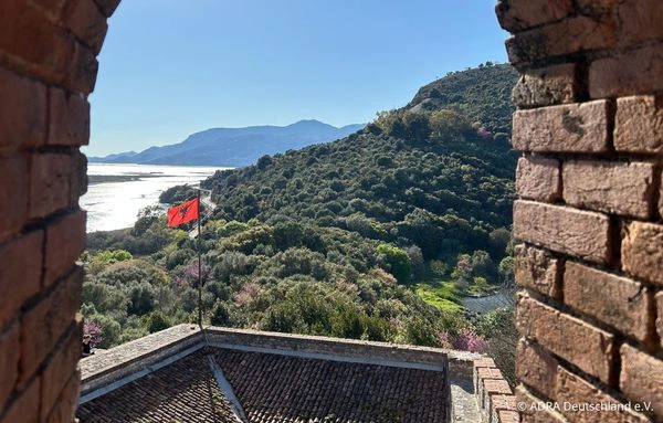 Ausblick durch einen steinernen Torbogen auf eine historische Anlage, grüne Hügel und eine albanische Flagge in der Ferne