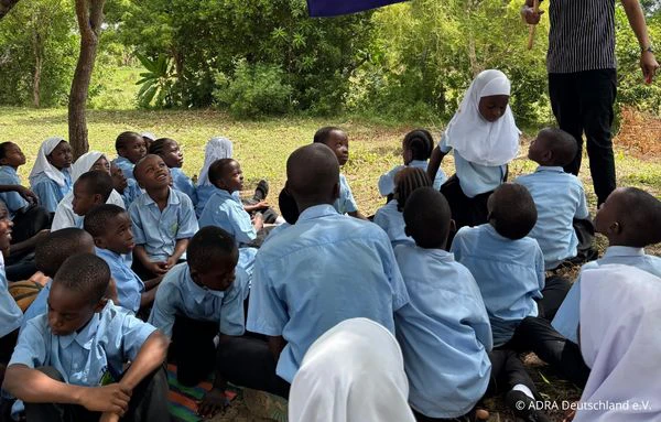 Schülerinnen und Schüler von The Baobab Home sitzen gemeinsam unter Bäumen, während ein Mitarbeiter eine große blaue Fahne hält