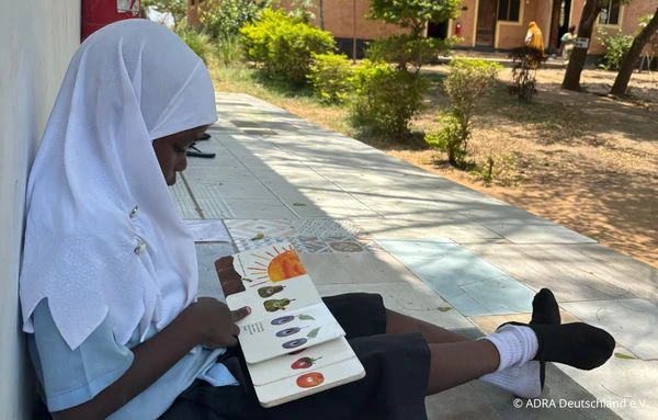 Schülerin sitzt auf dem Schulgelände von The Baobab Home und arbeitet mit einem bebilderten Lernmaterial