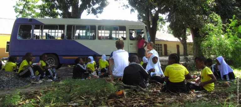 Schülerinnen und Schüler von The Baobab Home sitzen gemeinsam mit Freiwilligen im Schatten großer Bäume neben einem Schulbus