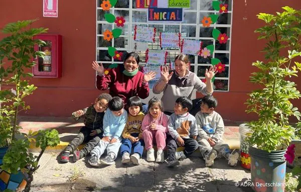 Eine ADRAlive!-Freiwillige und eine Betreuerin sitzen mit sechs Kindern vor der Kindertagesstätte Nuestro Hogar