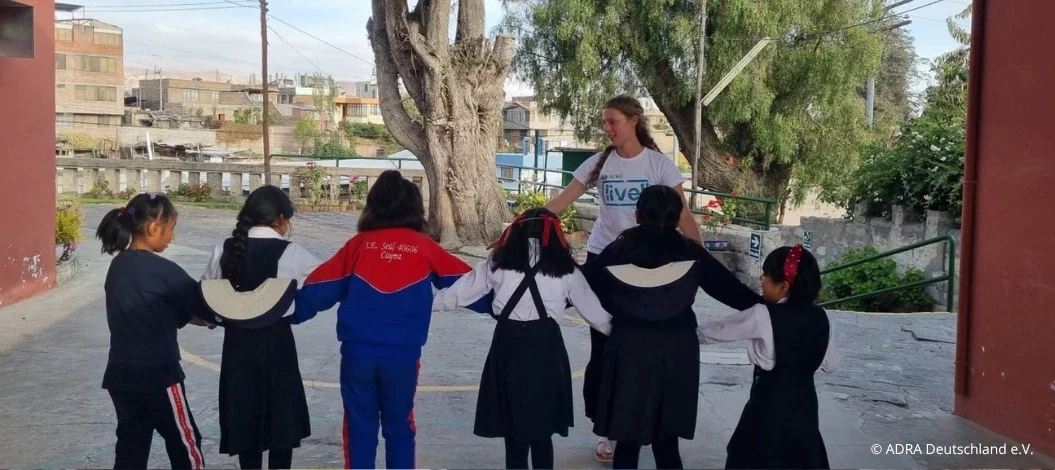 Header_Erfahrungsbericht_Sophie_in_Peru ADRAlive!-Freiwillige pielt in Peru, spielt mit einer Gruppe von Schulkindern, die sich im Kreis an den Händen halten, auf einem Schulhof.