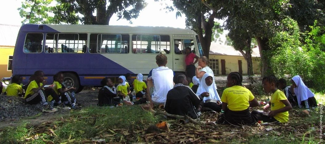 Schülerinnen und Schüler von The Baobab Home sitzen gemeinsam mit Freiwilligen im Schatten großer Bäume neben einem Schulbus